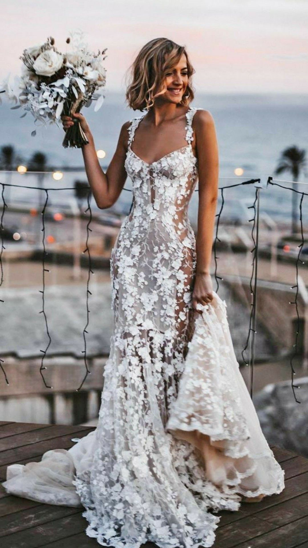 Woman in a white lace wedding dress holding a bouquet on a dock with palm trees in the background.