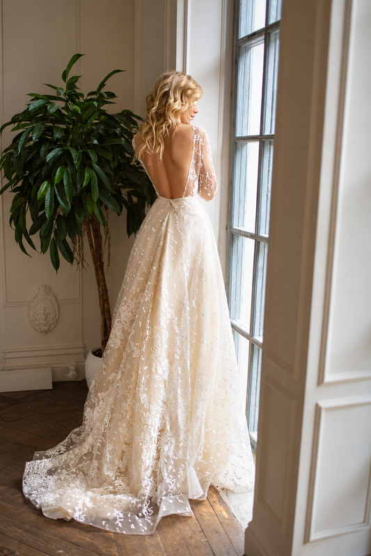 A woman wearing a voluminous ivory lace wedding gown with long sleeves and a fitted bodice, standing in a bright indoor room with plants and framed wall art. - HER SHOP®