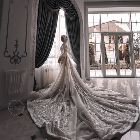 A woman wearing a long-sleeved white lace modest bridal gown with a matching sheer train and hijab, standing in a grand, arched hall with mirrors and draped fabrics in the background. - HER SHOP®