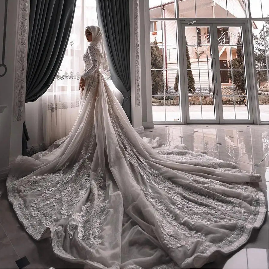 A woman wearing a long modest white lace bridal gown with a dramatic train stands in a bright room with tall windows and dark gray curtains, facing a window. The dress has long sleeves, a fitted silhouette, and an elaborate embroidered train spread across the floor.