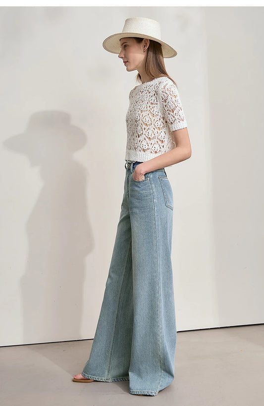 Woman wearing light blue jeans and a white lace top, holding a brown handbag on a plain background with AMII branding. - HER SHOP