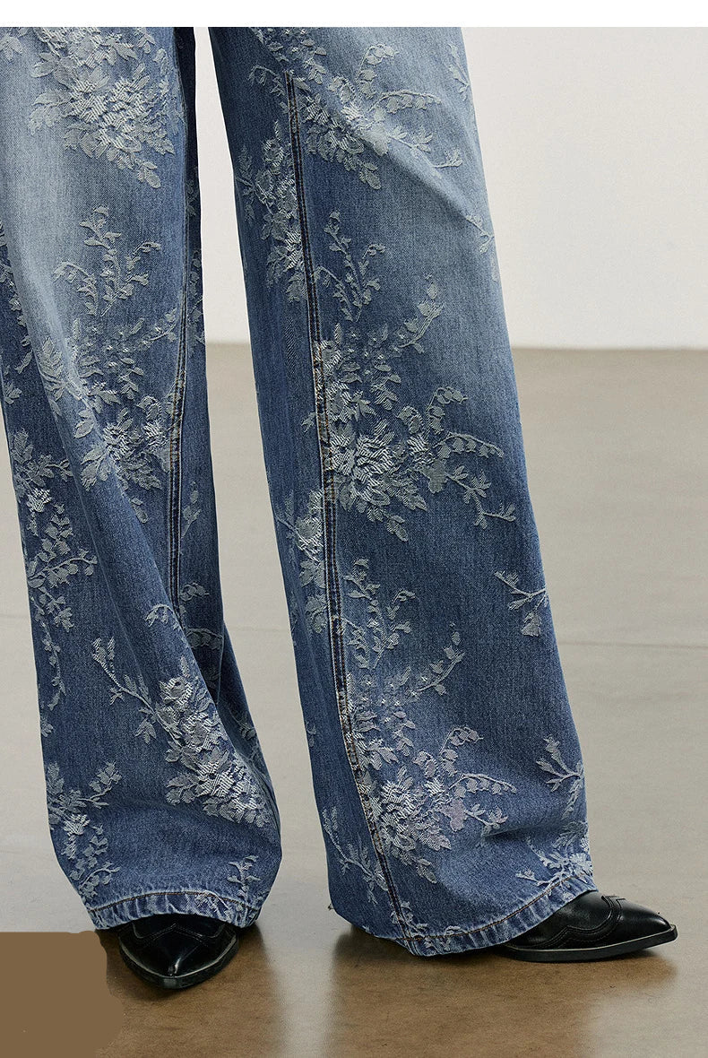 Blue jeans with floral embroidery worn by a person on a light wooden floor.