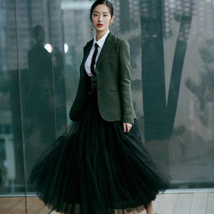 Woman in a green blazer and black tulle skirt standing in front of a glass wall.