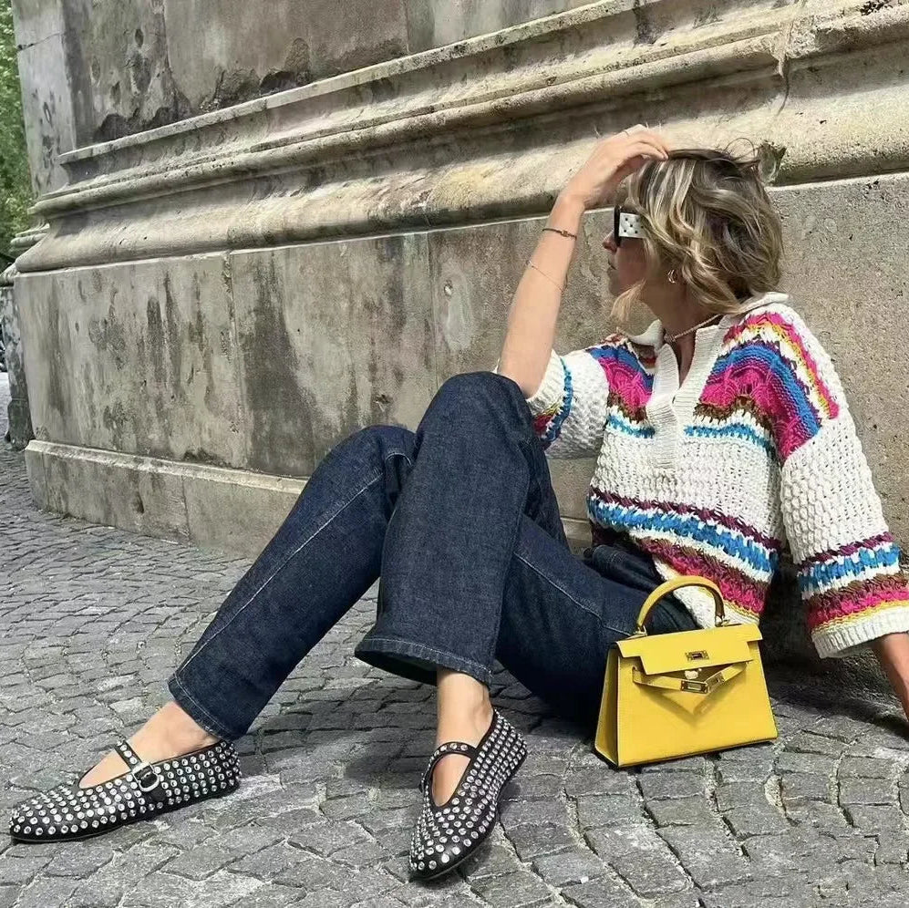 Woman sitting on a stone pavement wearing a colorful sweater and jeans, with a yellow handbag beside her, wearing flat shoes with square crystal