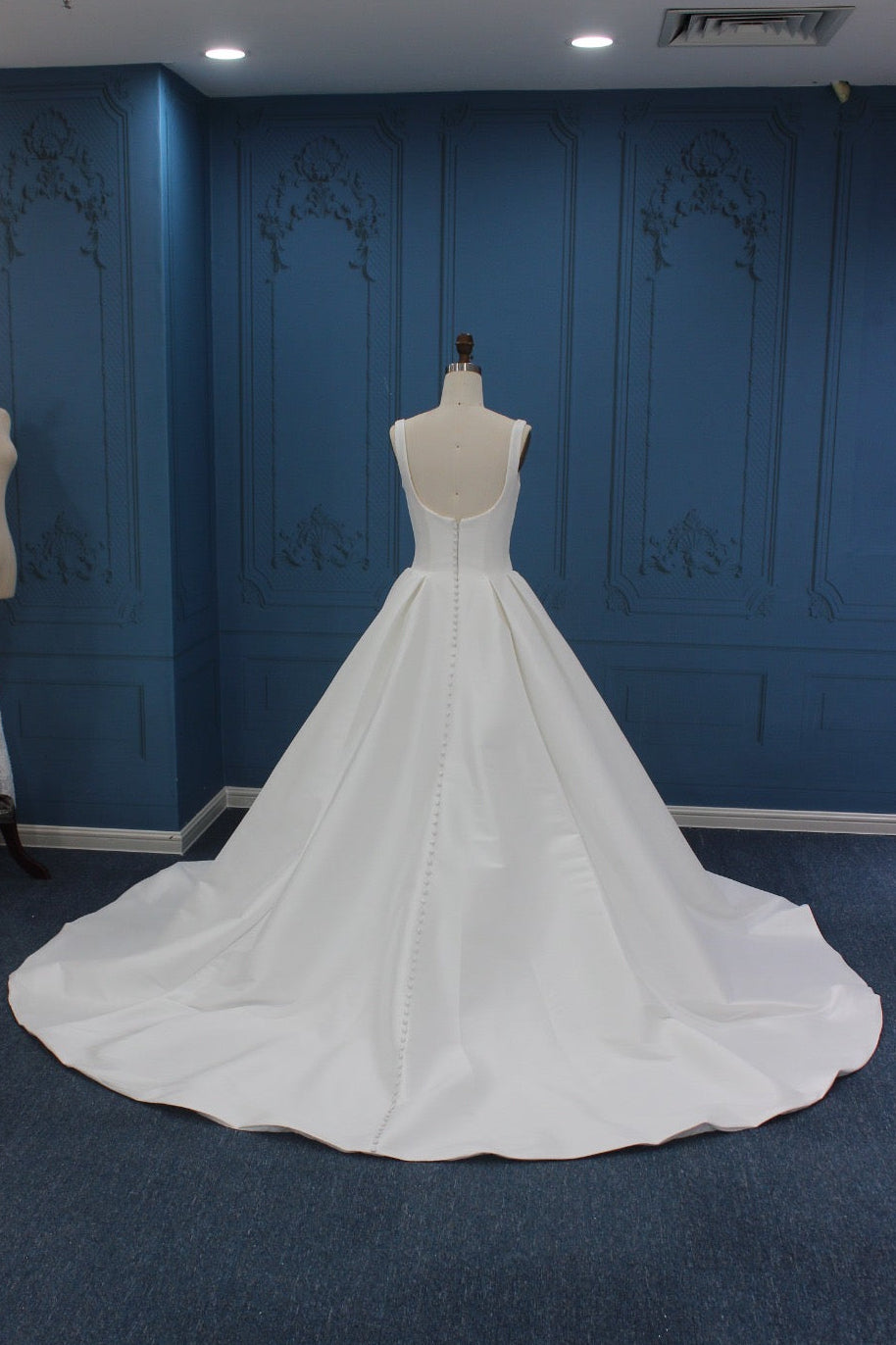 A white wedding dress displayed on a mannequin in a bridal boutique, featuring a sleeveless backless design with a long train on a blue carpeted floor and paneled blue wall.