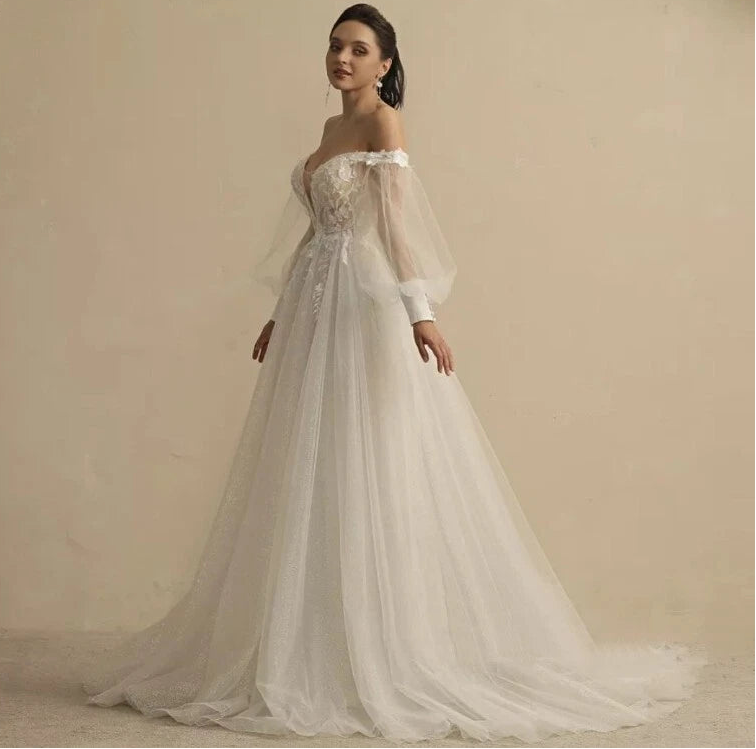 An off-the-shoulder A-line wedding dress with a lace bodice, sheer long sleeves, and a voluminous tulle skirt, shown on a model against a neutral beige background.