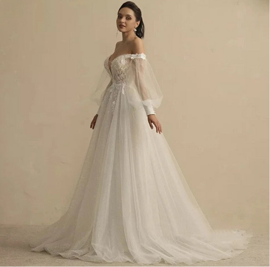 An off-the-shoulder A-line wedding dress with long sheer puff sleeves, a lace-embellished bodice, and a flowing tulle skirt, shown on a model against a warm beige studio background. - HER SHOP®