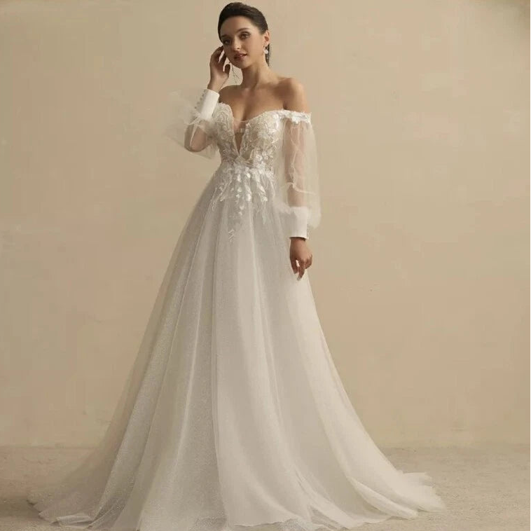 An off-the-shoulder A-line wedding dress with long sheer puff sleeves, a lace-embellished bodice, and a flowing tulle skirt, shown on a model against a warm beige studio background.