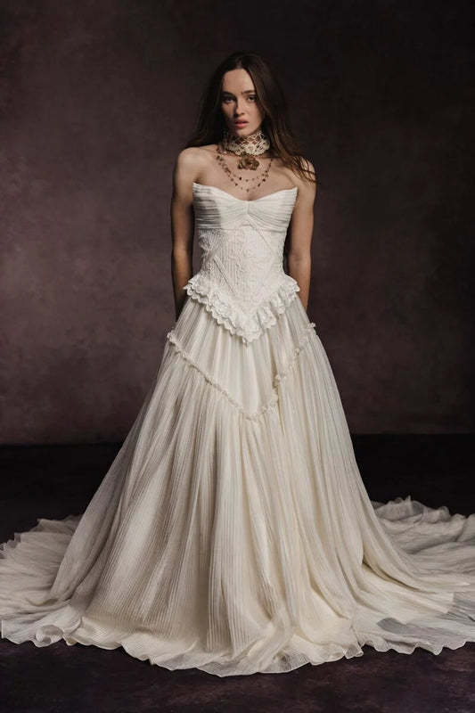 A woman wearing a strapless ivory bridal gown with a structured lace bodice, ruffled waist, and a voluminous pleated skirt, posed against a dark brown studio background. - HER SHOP®