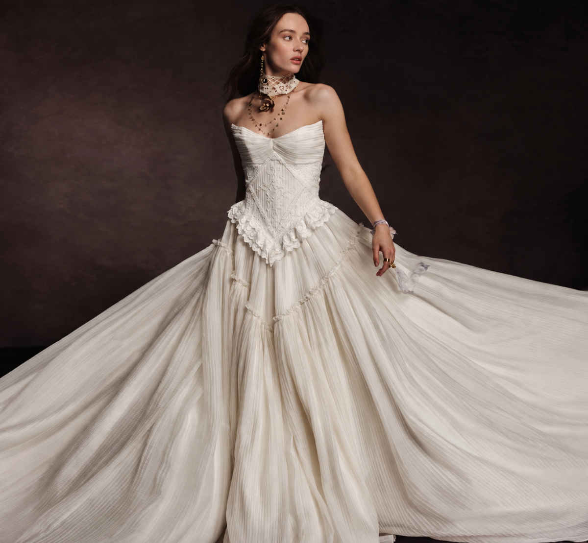 A woman wearing a strapless ivory bridal gown with a structured lace bodice, ruffled waist, and a voluminous pleated skirt, posed against a dark brown studio background. - HER SHOP®