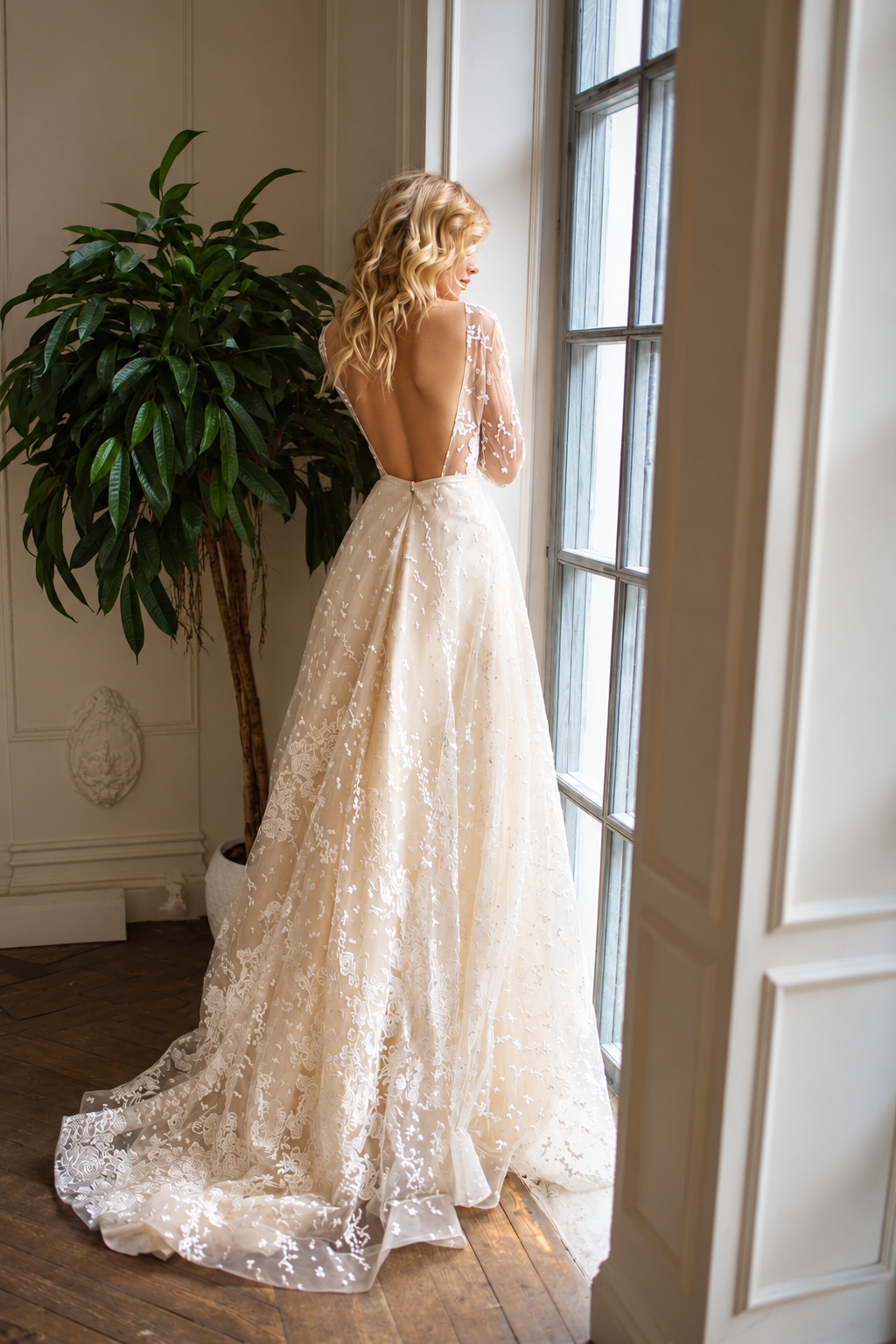 A woman stands by a window wearing a long-sleeved lace wedding gown with an open back and a full train, photographed in a bright indoor room with a potted plant and wood floor.