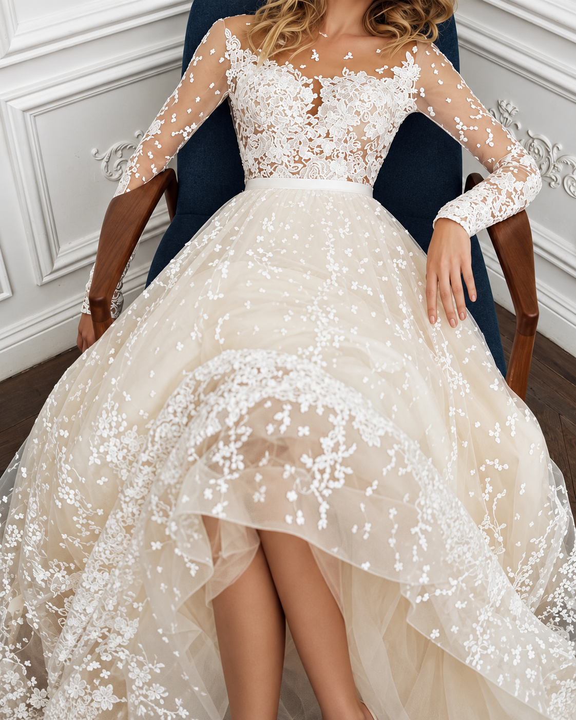 A woman seated in a dark blue armchair wearing a voluminous ivory wedding gown with sheer long sleeves, a lace bodice, and a layered tulle overskirt with floral applique.
