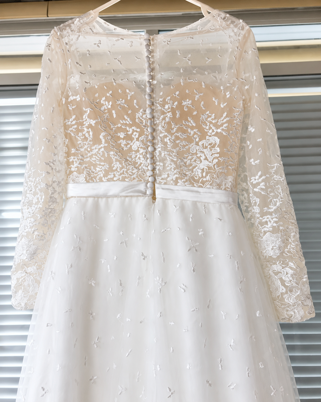 A white and champagne lace wedding dress with long sleeves and a beaded button-back detail, hanging against a window blind background.