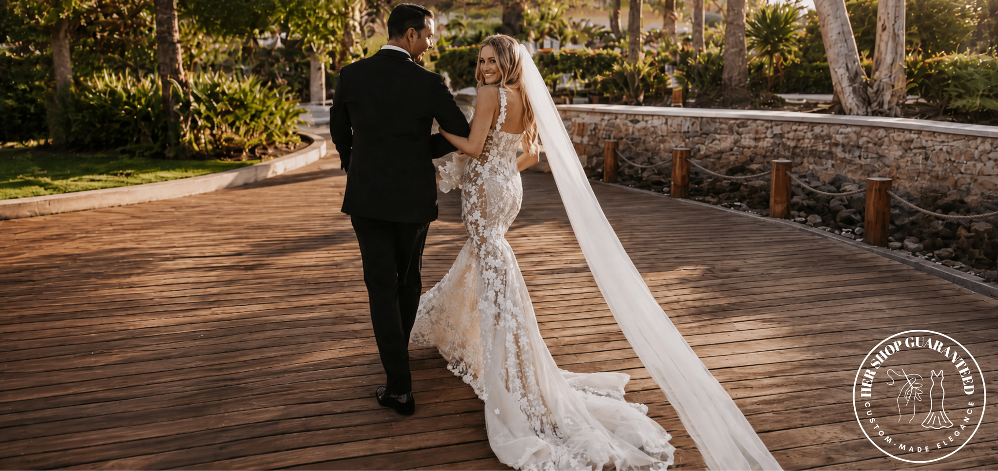 A bride in a sleeveless lace mermaid wedding gown with a long sheer veil walks hand in hand with a groom in a black suit along a curved wooden boardwalk outdoors. Trees, plants, and a stone wall are visible in the background.
