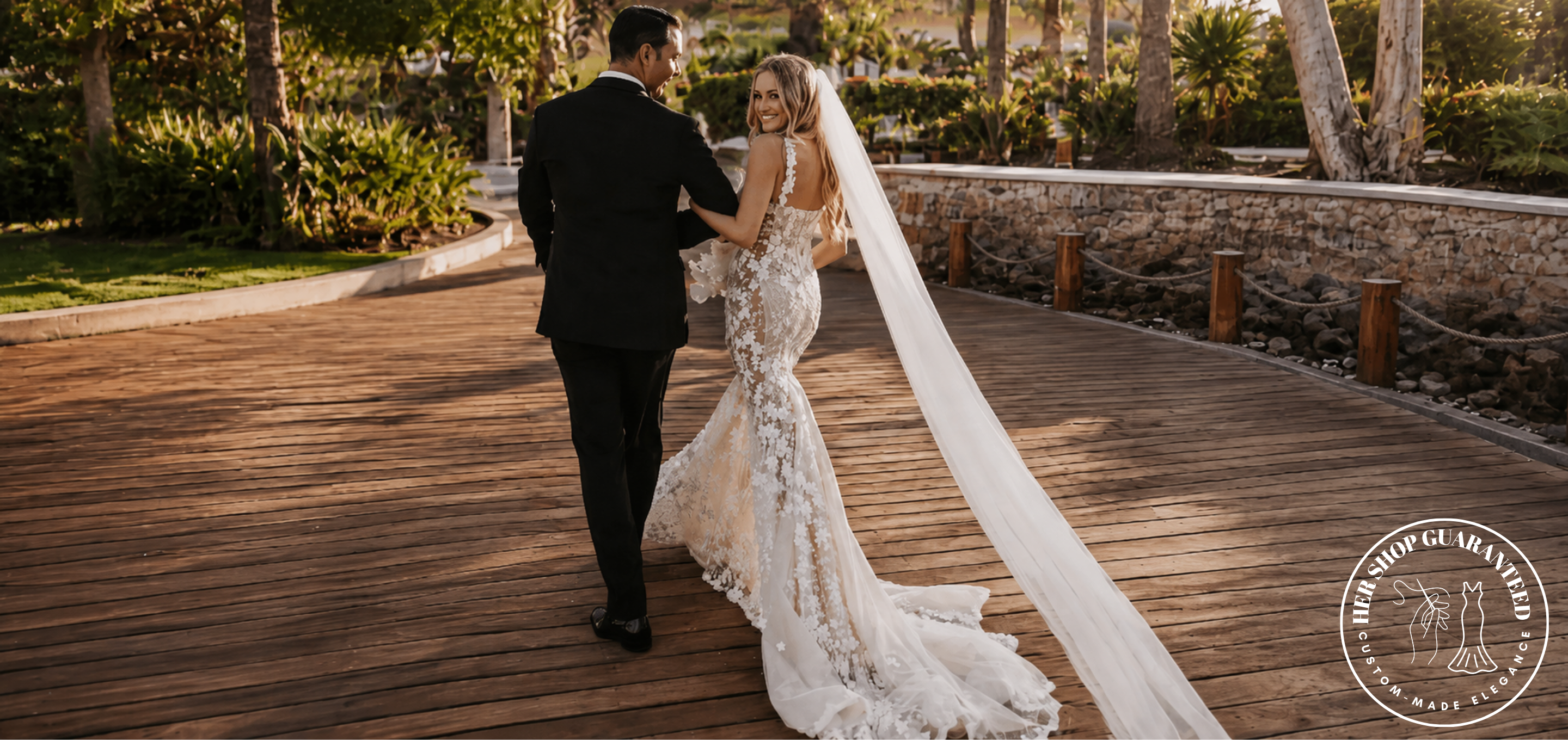 A bride in a sleeveless lace mermaid wedding gown with a long sheer veil walks hand in hand with a groom in a black suit along a curved wooden boardwalk outdoors. Trees, plants, and a stone wall are visible in the background.