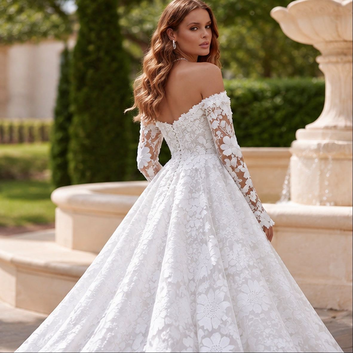 A woman wearing a white off-the-shoulder lace wedding gown with long sheer sleeves and a full ball gown skirt, photographed outdoors near a stone fountain and greenery.