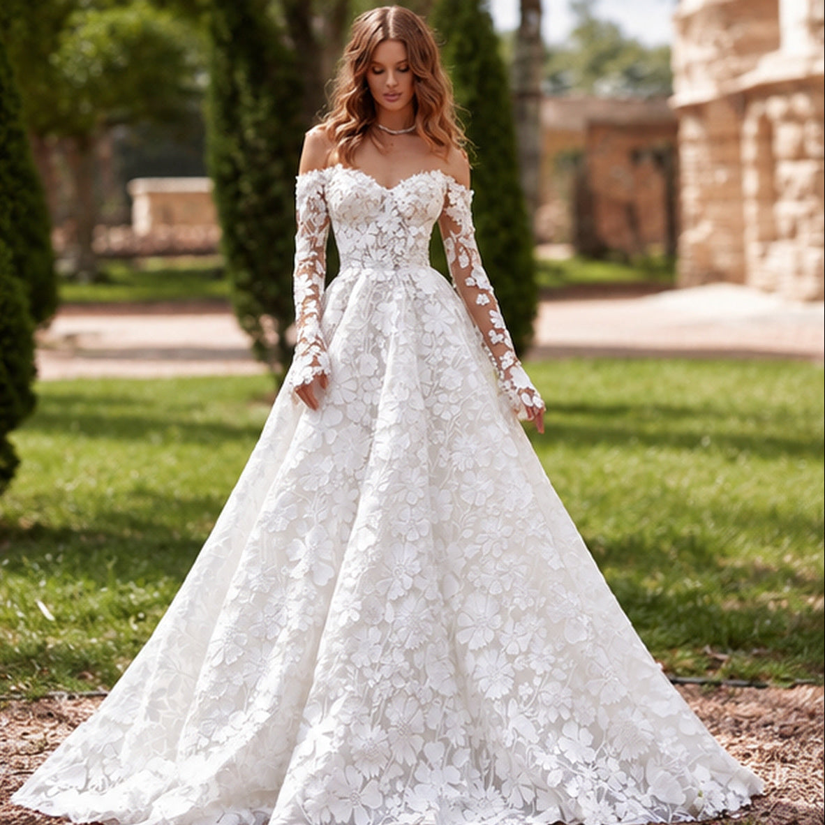 A woman wearing a white off-the-shoulder lace wedding gown with long sheer sleeves and a full A-line skirt, standing outdoors on grass with trees and a stone building blurred in the background.
