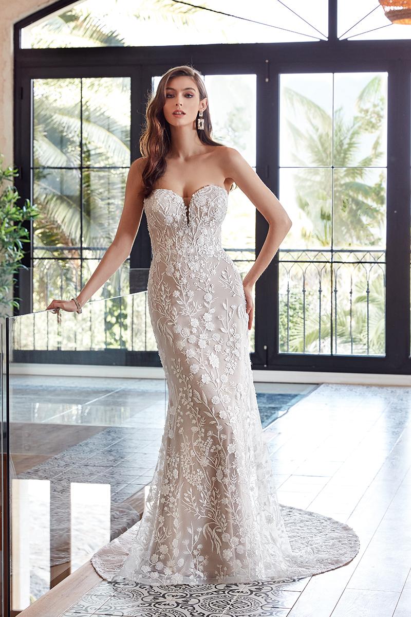 A woman models a strapless ivory lace mermaid-style wedding gown with a sweetheart neckline and floral applique, standing in a bright interior with large windows and palm trees visible outside.