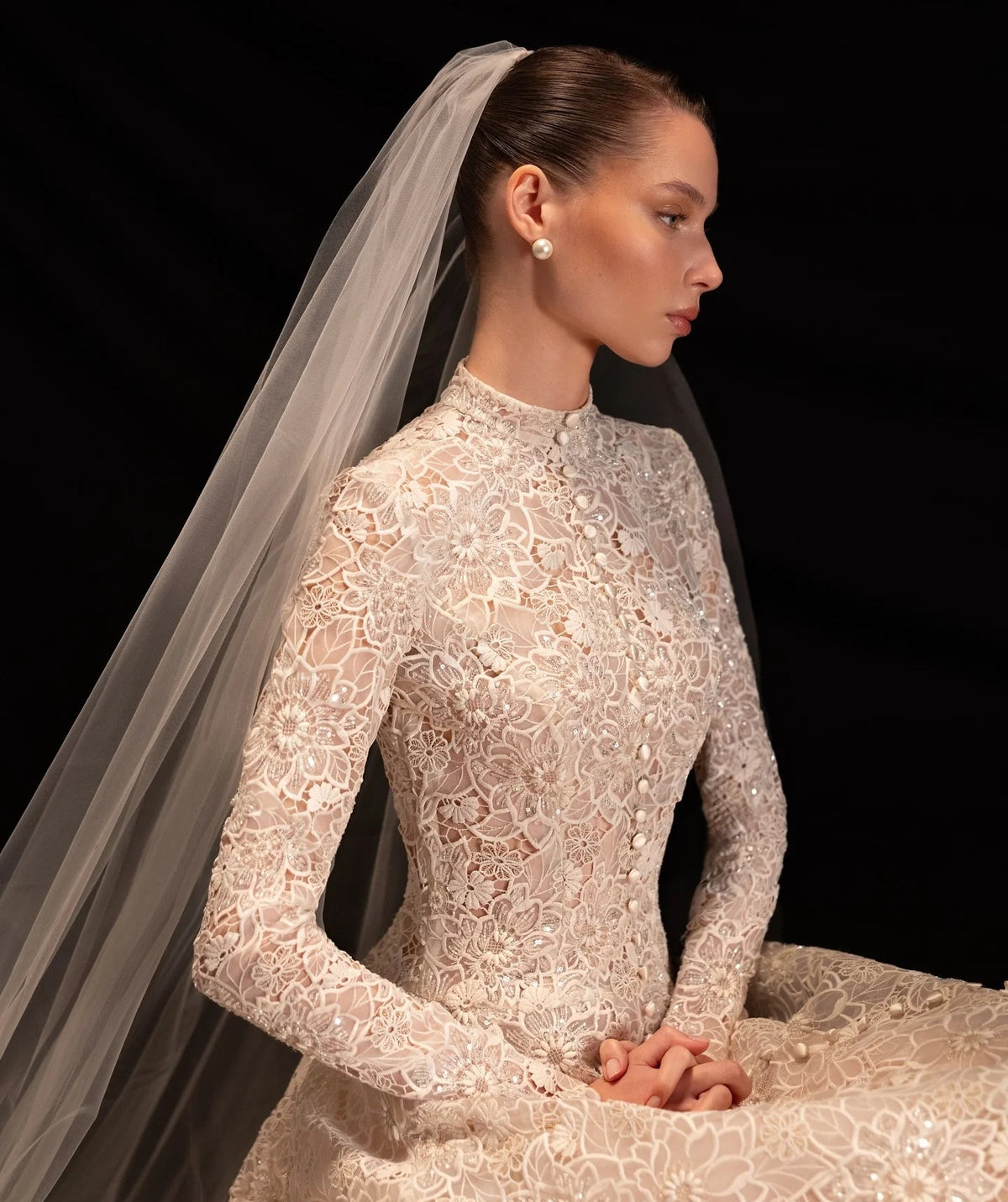 A woman in profile wearing a long-sleeve lace modest bridal gown with a sheer veil, posed against a black background. The dress has an intricate floral lace pattern and a high neckline.