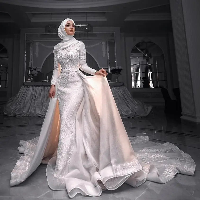 A woman wearing a long-sleeved white lace modest bridal gown with a matching sheer train and hijab, standing in a grand, arched hall with mirrors and draped fabrics in the background.