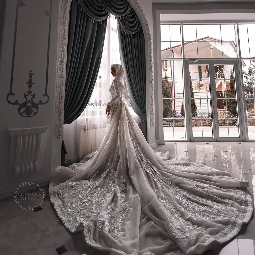 A woman wearing a long modest white lace bridal gown with a dramatic train stands in a bright room with tall windows and dark gray curtains, facing a window. The dress has long sleeves, a fitted silhouette, and an elaborate embroidered train spread across the floor.