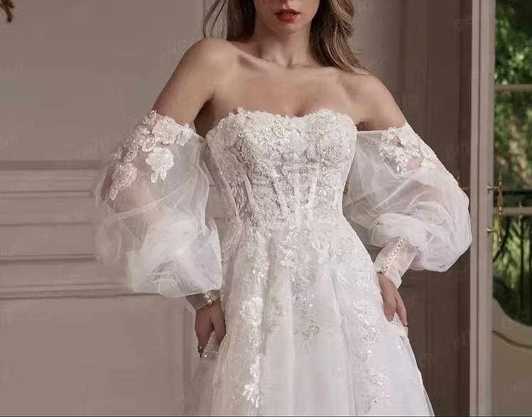 A woman wearing an off-the-shoulder white wedding gown with a fitted lace bodice, sheer balloon sleeves, and a full skirt, photographed indoors near a doorway.