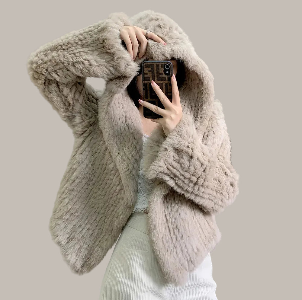 Person wearing a fluffy beige coat taking a mirror selfie in a room with a painting on the wall.