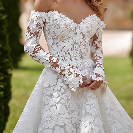 A woman wearing a white off-the-shoulder lace wedding gown with long sheer sleeves and a full A-line skirt, standing outdoors on grass with trees and a stone building blurred in the background. - HER SHOP®