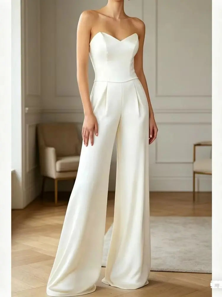 A woman modeling a strapless white jumpsuit with a sweetheart neckline, fitted bodice, and wide legs in an elegant indoor room with paneled walls and light wood flooring.