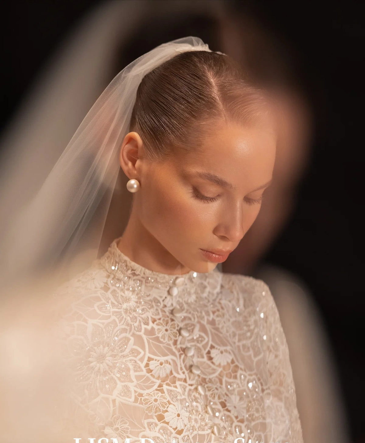 A close-up of a woman wearing a white lace modest bridal gown with a sheer veil and pearl earrings, photographed in warm, soft lighting against a dark background. The text 'LISM Dresses Store' appears at the bottom.