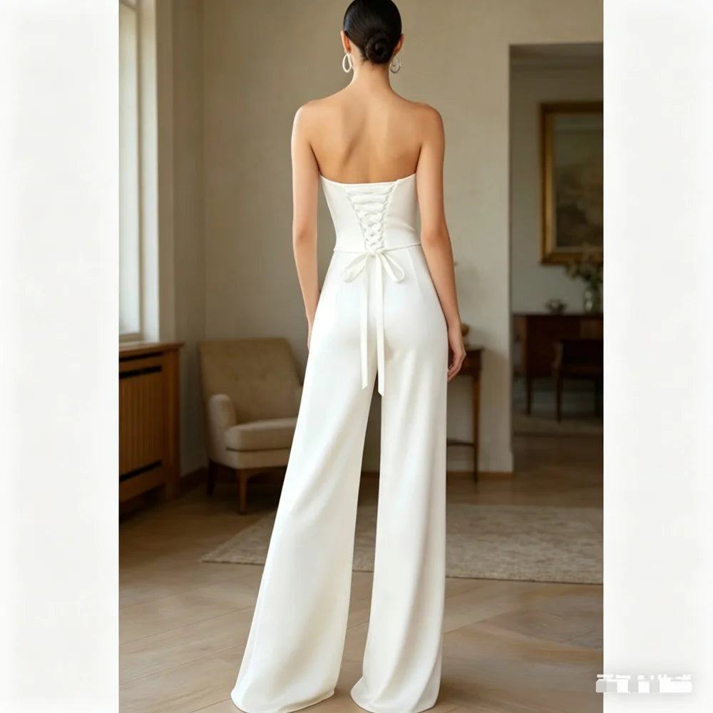 A woman stands with her back to the camera wearing a strapless white jumpsuit with a lace-up back and wide legs in a softly lit indoor room.