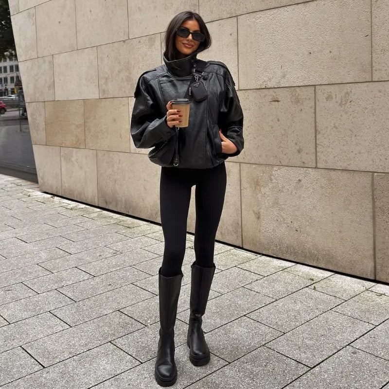 Person wearing a black outfit with a jacket, leggings, and boots, holding a coffee cup, standing against a stone wall.