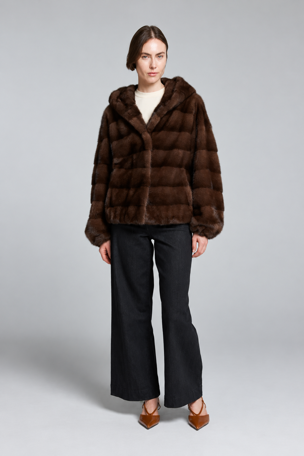 A model stands against a light gray studio background wearing a dark brown plush faux fur jacket with a hood, black wide-leg trousers, and brown pointed-toe heels.