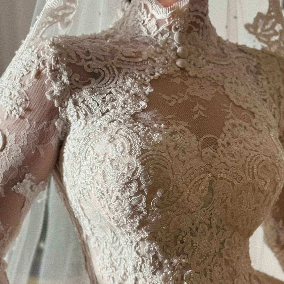 Close-up of a lace wedding dress with sheer long sleeves, floral embroidery, and a high neckline, shown on a model. A watermark reading 'LIYNIC' is visible near the bottom.